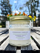 Load image into Gallery viewer, A soy candle with a label reading 'Rosemary Sage &amp; Citronella' placed on a wooden surface with a blurred background.
